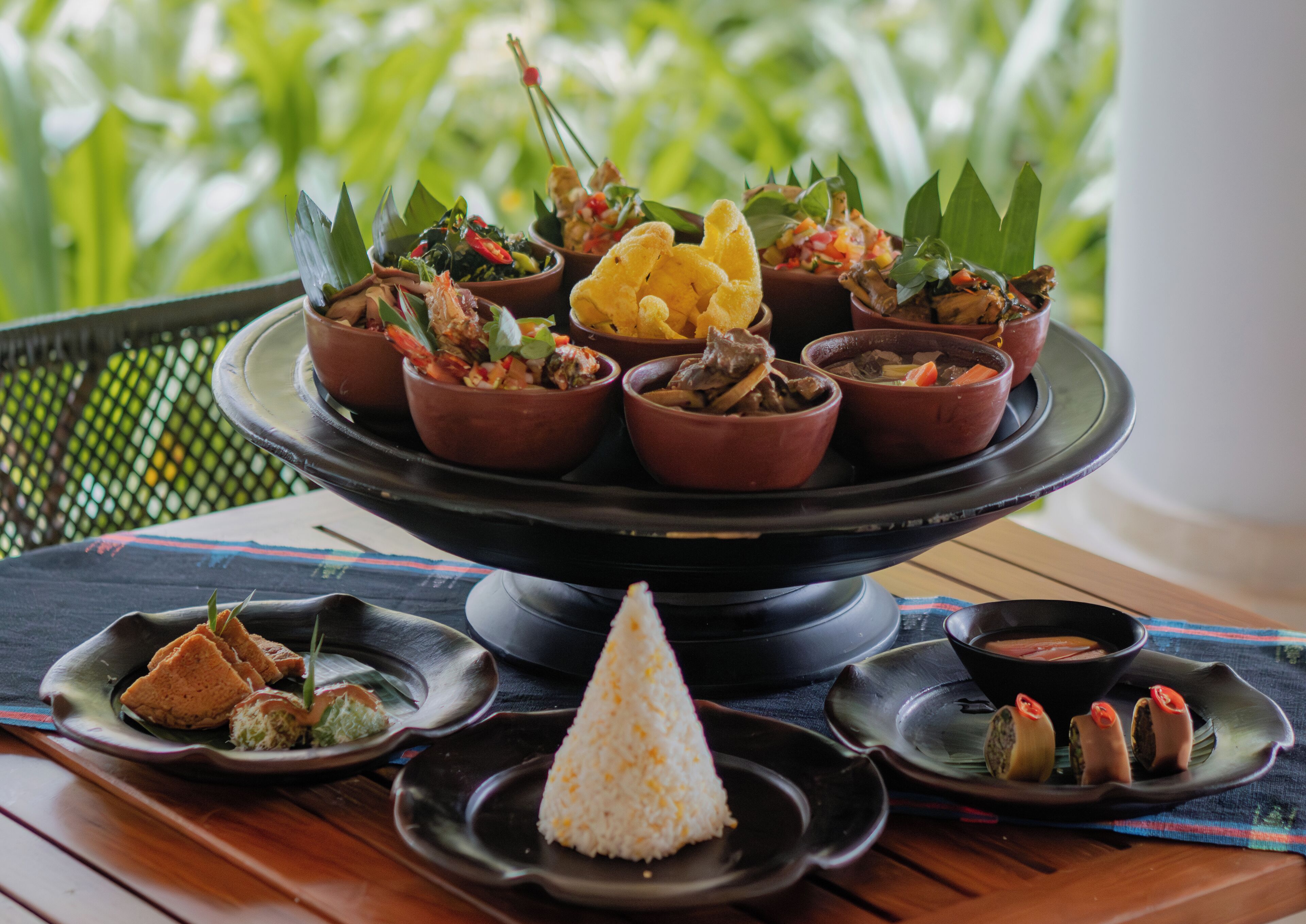 A multi-tiered serving dish with various dishes, including rice, vegetables, and meat, on a table with a view of greenery.