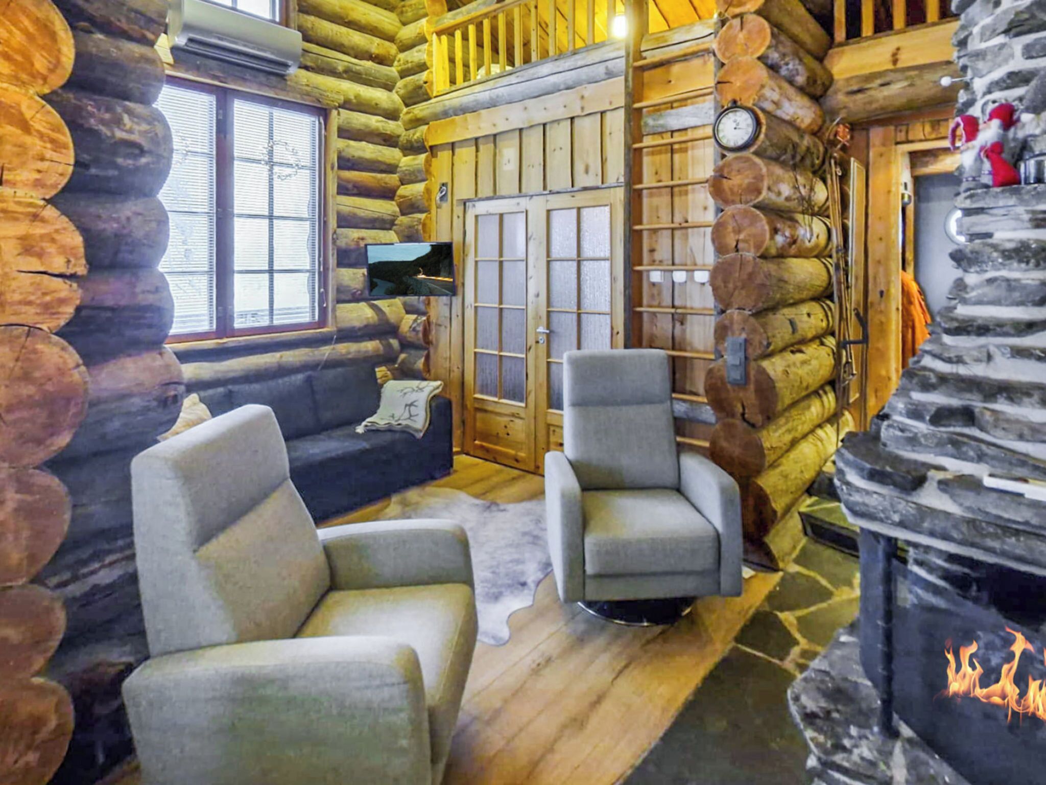 Ein Blockhaus-Interieur mit Kamin aus Stein, einer Couch, zwei Sesseln und einem großen Fenster.