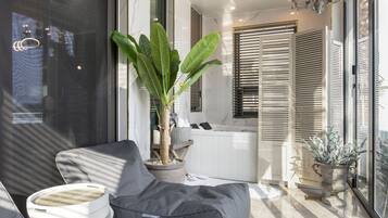 Relax in a serene space with a comfortable sofa, side table, and potted plant, featuring a bathtub and large windows with blinds.