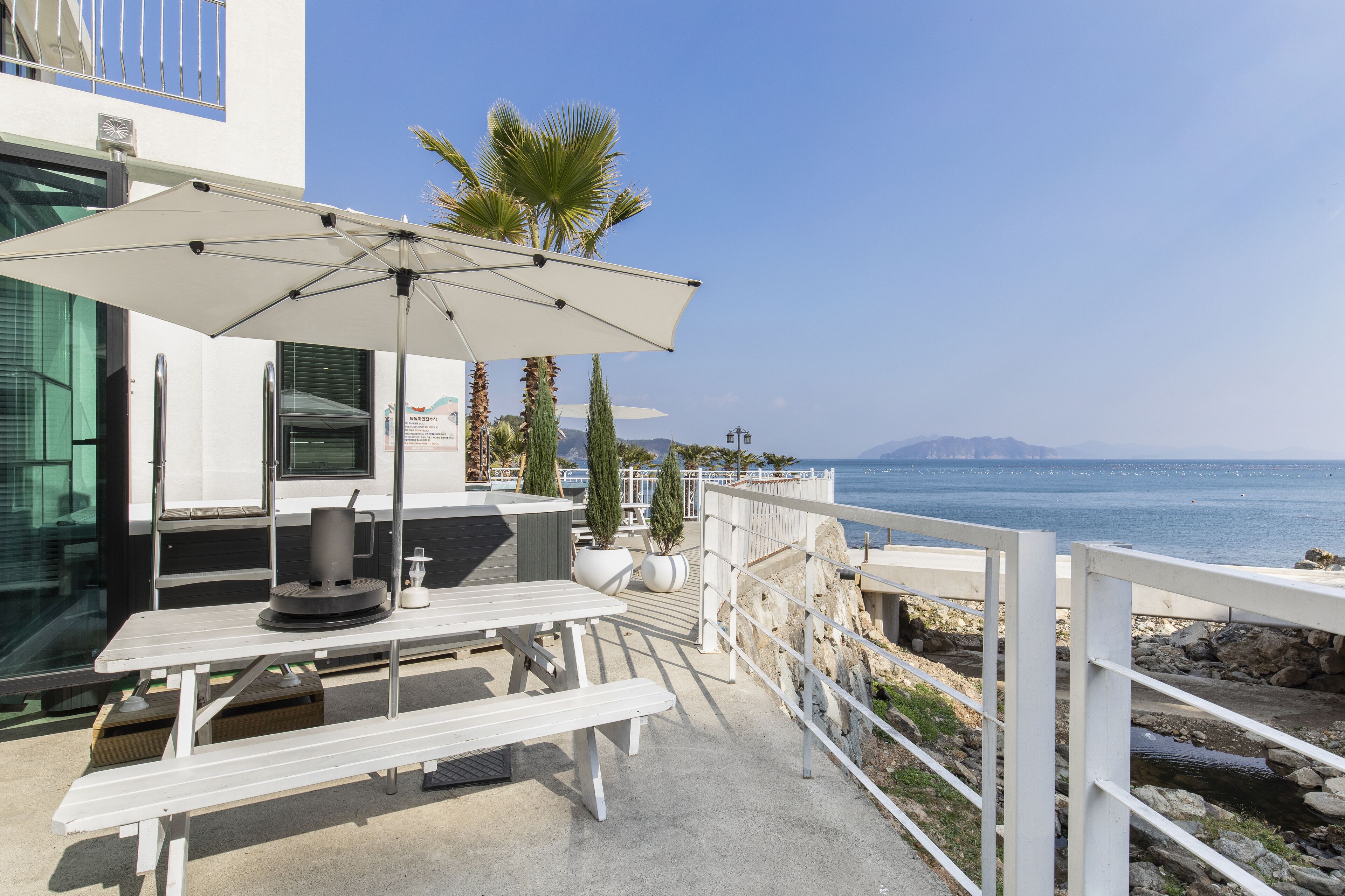 Una terraza frente al mar con una mesa y banca blancos, un paraguas grande, y vistas al mar y a las montañas.