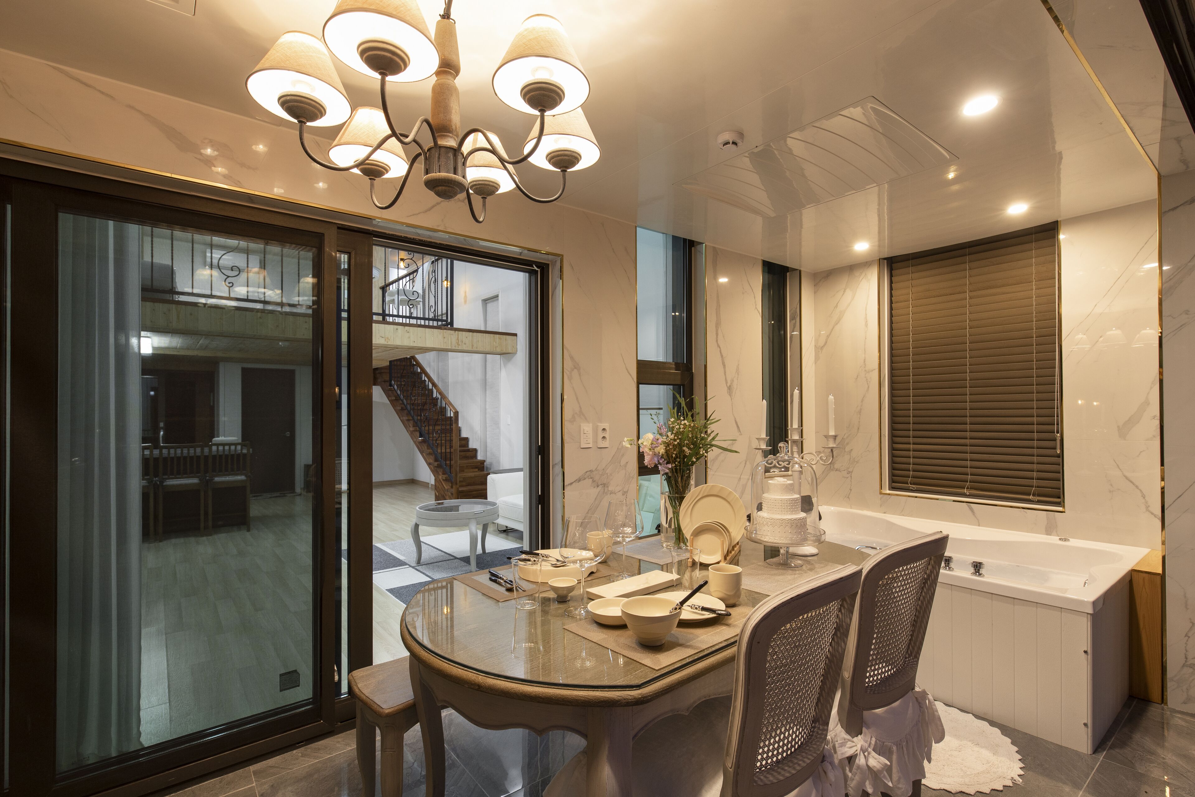 A modern dining area with a glass table, chairs, and a chandelier. A staircase is visible in the background leading to an upper level.