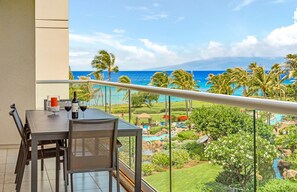 Un balcon avec une table dressée pour deux personnes, donnant sur une piscine et un paysage tropical avec des palmiers, et offrant une vue sur l’océan.