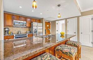 A kitchen with a central island, stainless steel appliances, wooden cabinets, and a backsplash. - Maui Resort Rentals: Honua Kai Hokulani 448 - Spacious 2BR w/ Partial Ocean & Mountain Views! (Lahaina)