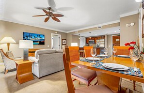 Une chambre d’hôtel avec un coin repas, un canapé, un ventilateur de plafond et une cuisine en arrière-plan.