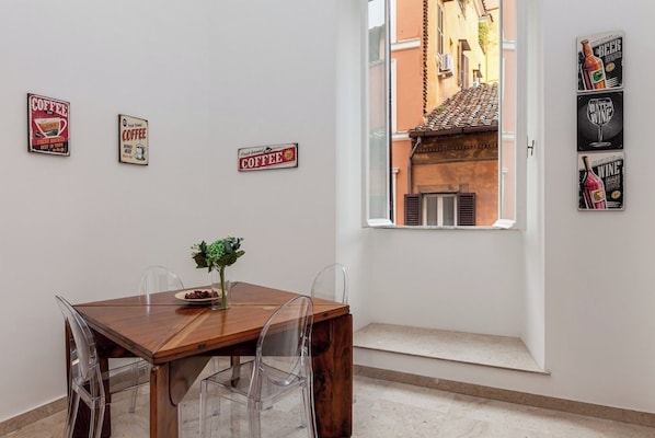 A dining area with a wooden table and clear chairs, a vase with flowers, and a window with a view of a building.