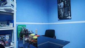 Een vergaderruimte met een blauwe muur, een glazen tafel en een zwarte kantoorstoel. Links staan schappen met dozen en een groen elektronisch apparaat.