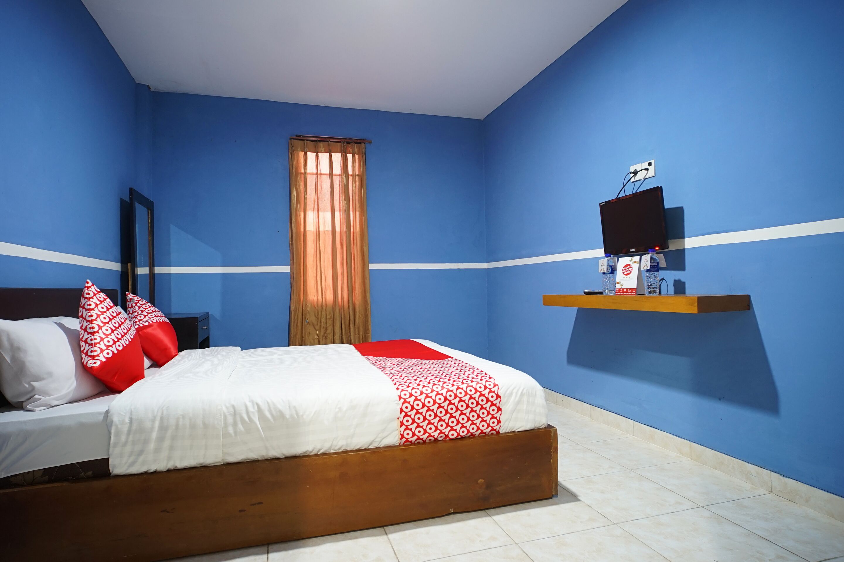 Une chambre à coucher avec un lit, une fenêtre avec des rideaux, un téléviseur fixé au mur et une étagère avec divers objets.