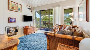 A spacious living room with a large TV, wooden furniture, and a view of the outdoors through sliding glass doors.