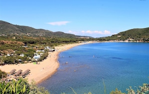 A coastal beach with clear blue water, sandy shore, and surrounding hills.