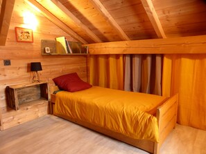 A cozy wooden cabin room with a single bed, a nightstand, a lamp, and a shelf with books and a framed picture.