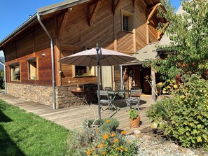 A wooden cabin with a patio area featuring a table and chairs, a small kitchen, and a garden with flowers and plants.