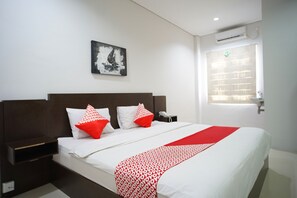 A hotel room with a large bed, white bedding, red and white pillows, a red and white patterned throw, a nightstand, a phone, a wall-mounted air conditioner, and a window with blinds.