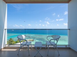 Varanda com duas cadeiras brancas e uma mesa pequena, com vista para o mar azul e límpido onde se vê um barco e um paraquedas a certa distância.