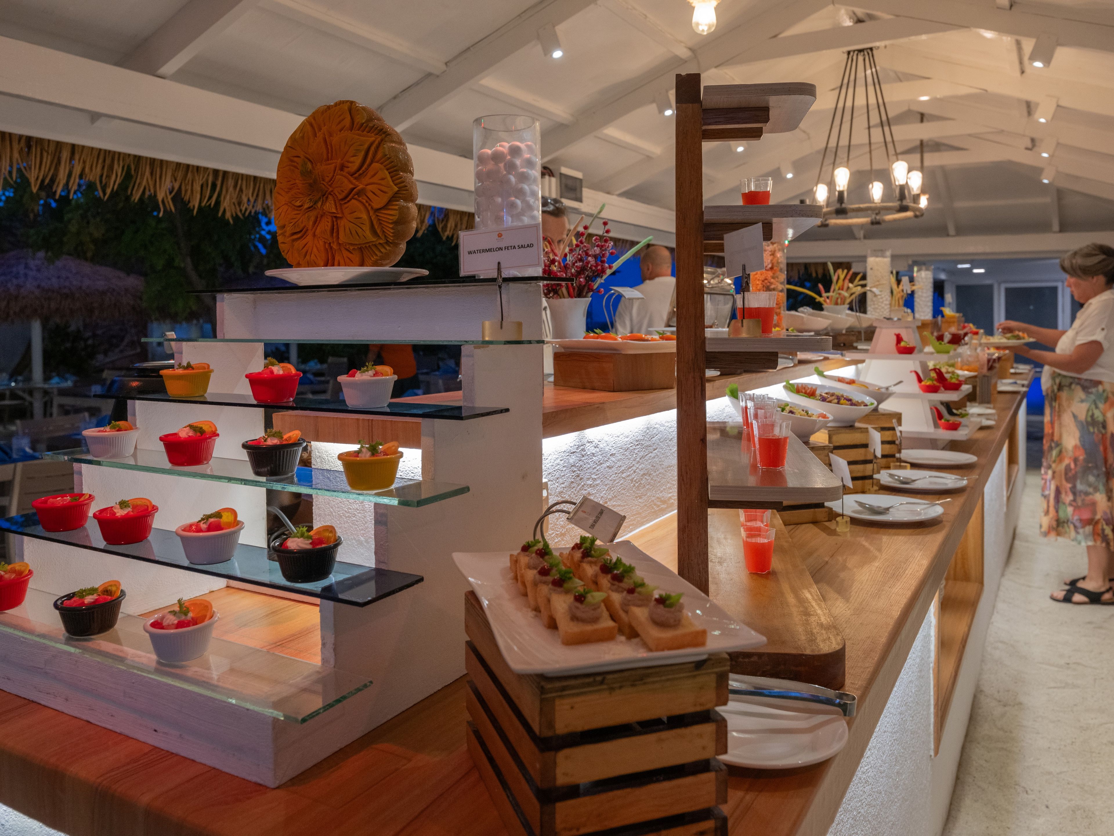 Un buffet proposant divers plats et une pancarte indiquant la présence d’une salade de pastèque et de feta.