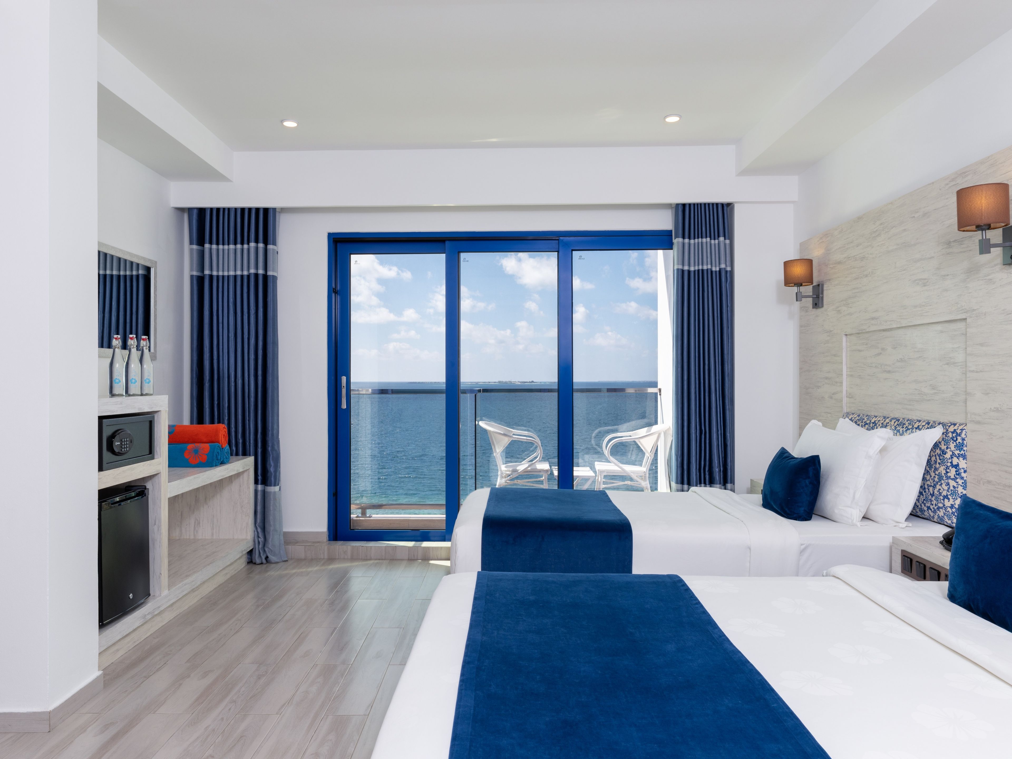 Habitación de hotel con un amplio ventanal corredizo al mar, una cama con ropa de cama azul, un pequeño escritorio con lámpara y un minibar.