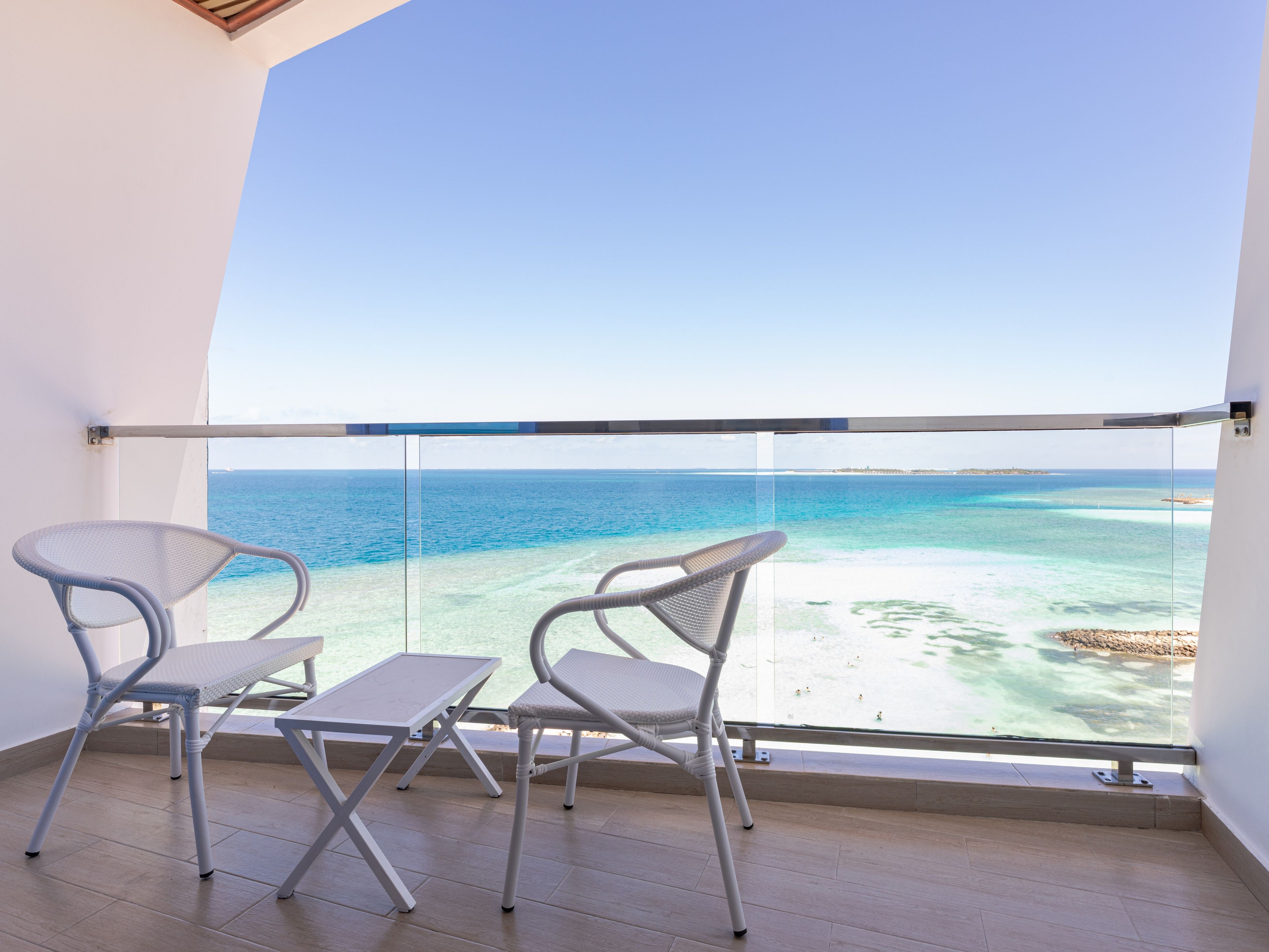 A balcony with two chairs and a table, overlooking a beach with clear blue water and a distant shoreline.