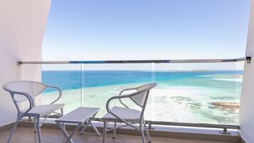 Un balcón con dos sillas y una mesa, con vista a una playa de aguas azules cristalinas y una costa lejana.