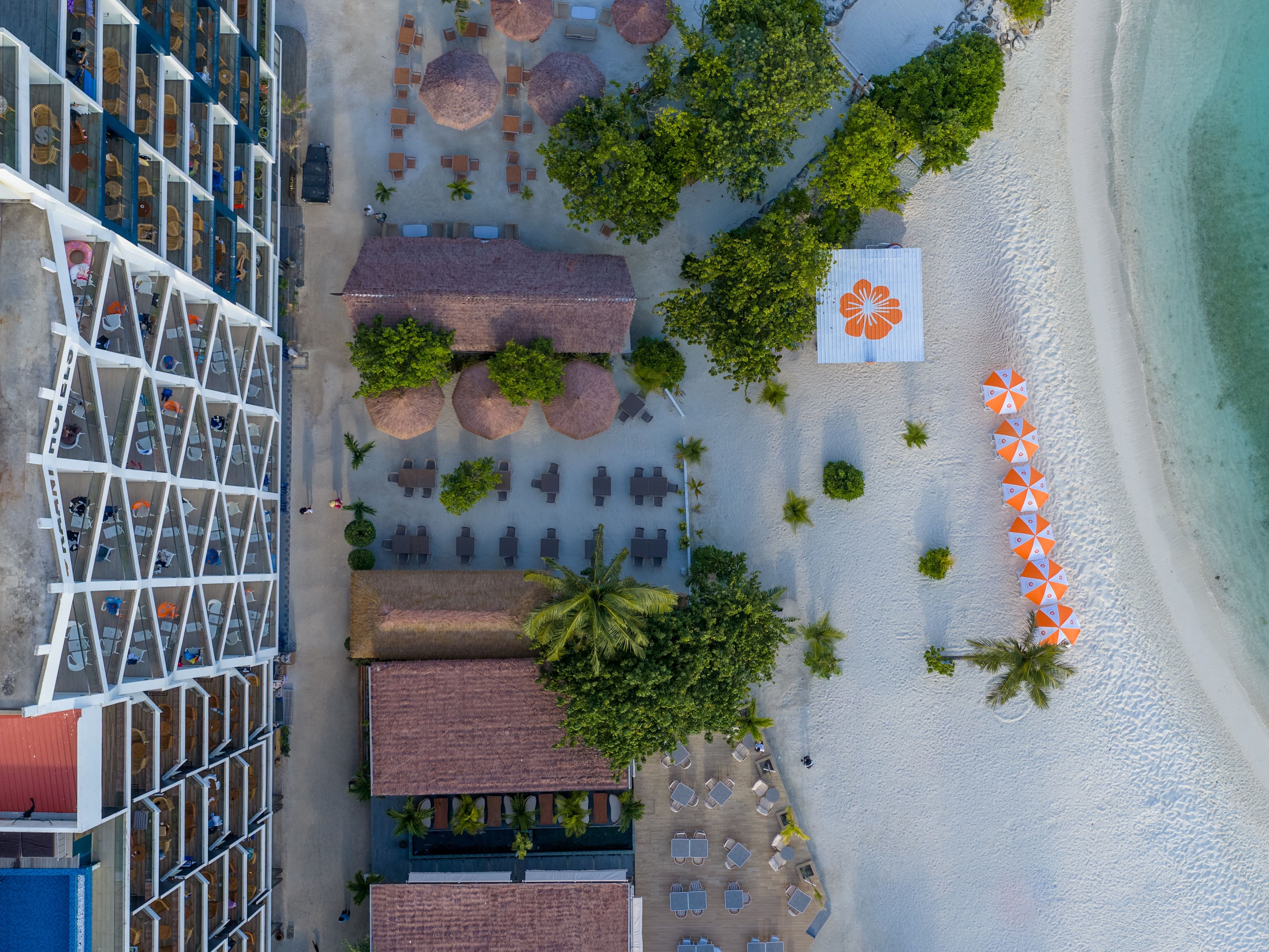 Vista aérea de un hotel frente a la playa con varias zonas de comedor, asientos al aire libre y un gran paraguas.