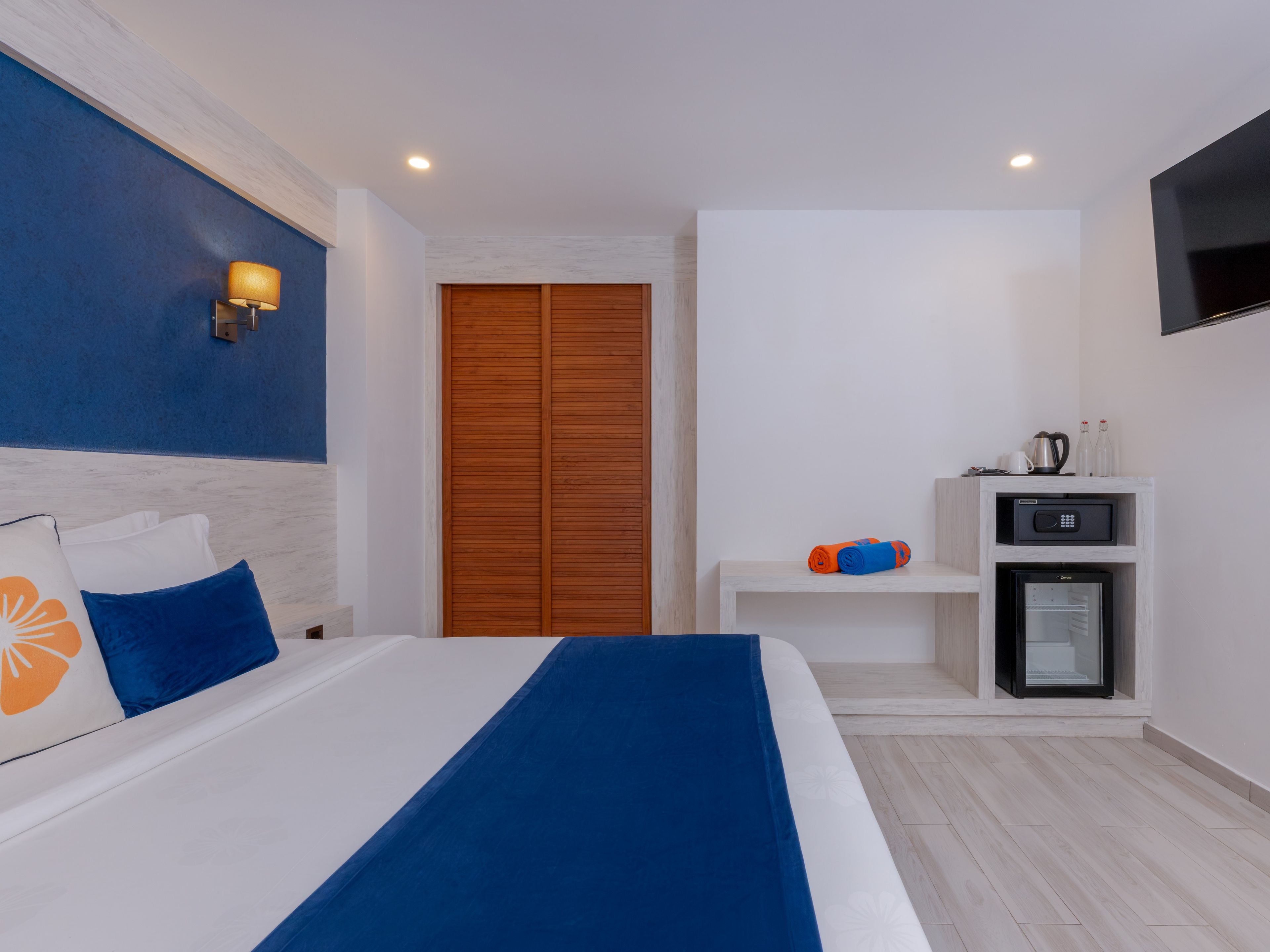 A hotel room with a bed, a wardrobe, a TV, a mini-fridge, and a shelf with toiletries.