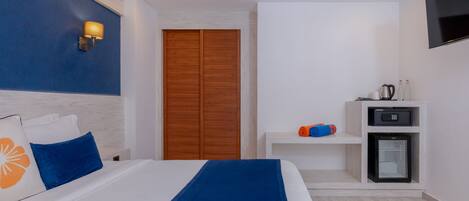 Habitación de hotel con cama, armario, televisión, mini refrigerador y estante con artículos de aseo.