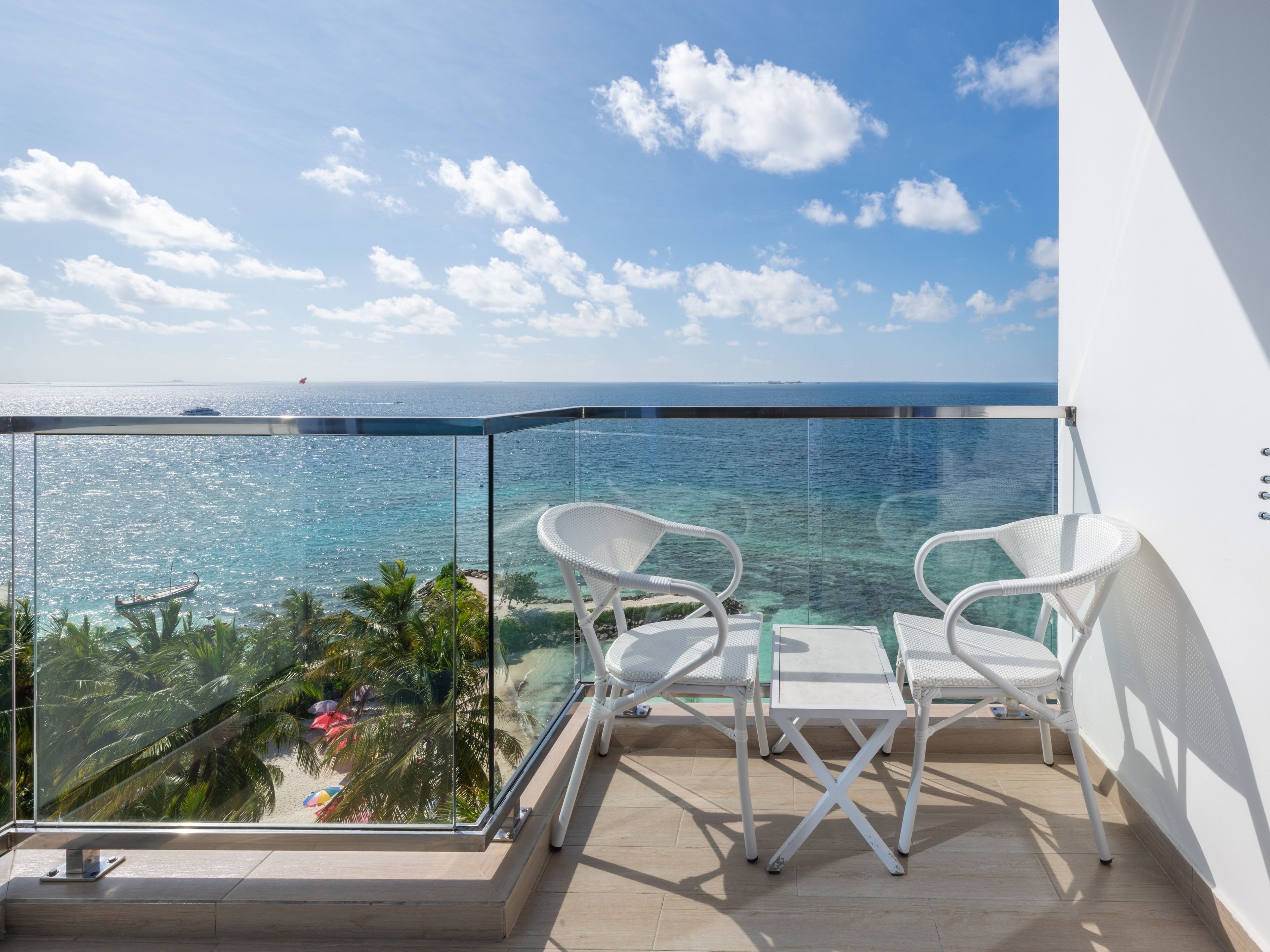 A balcony with a glass railing overlooking a beach and the sea. There are two white chairs and a small table.