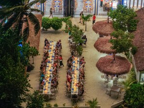 Un área de comedor al aire libre con mesas y sillas, rodeada de palmeras y sombrillas de paja.