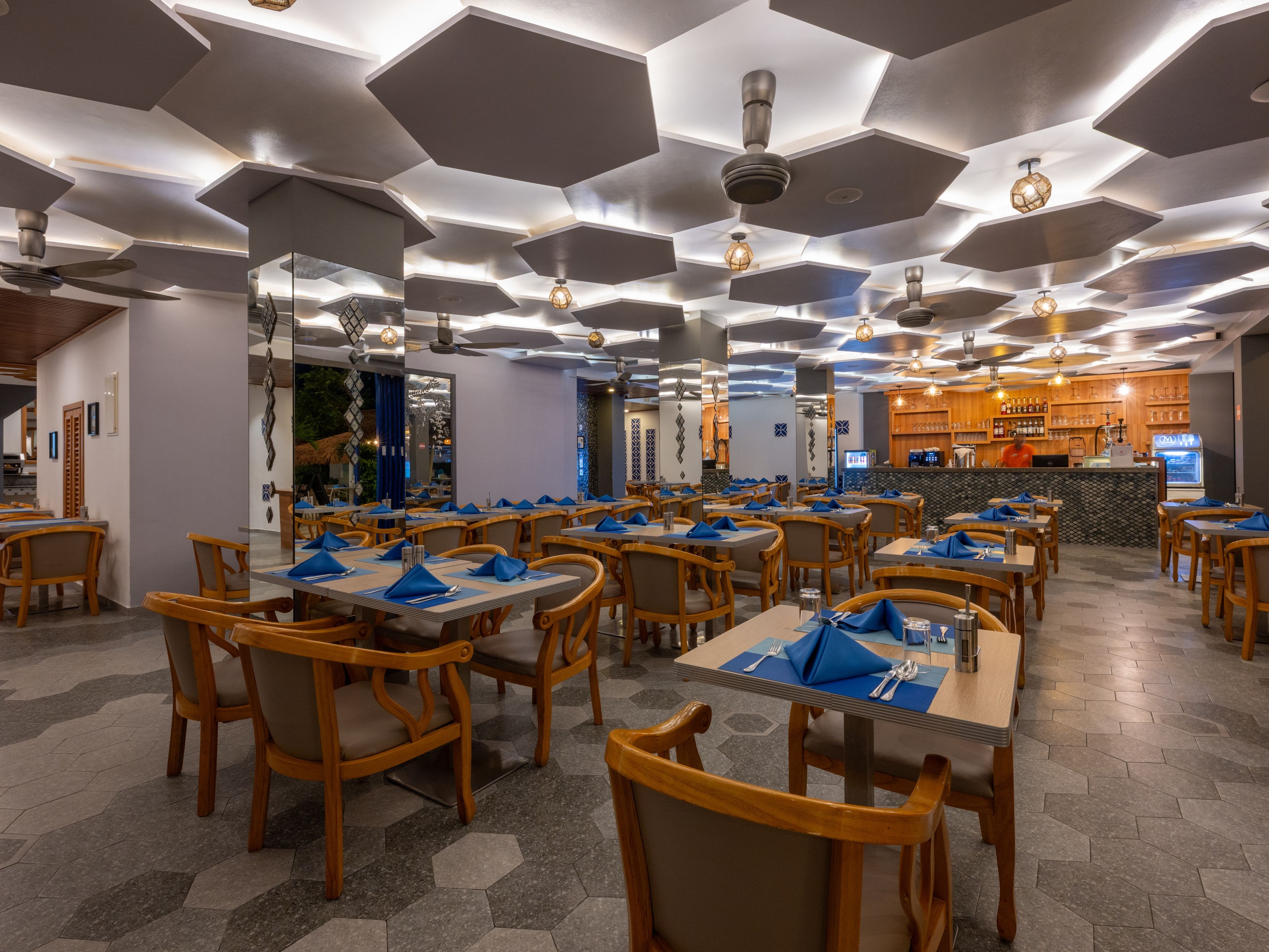 Ein Restaurant mit sechseckigen Deckenplatten, Holztischen mit blauen Servietten und einer Bar im Hintergrund.