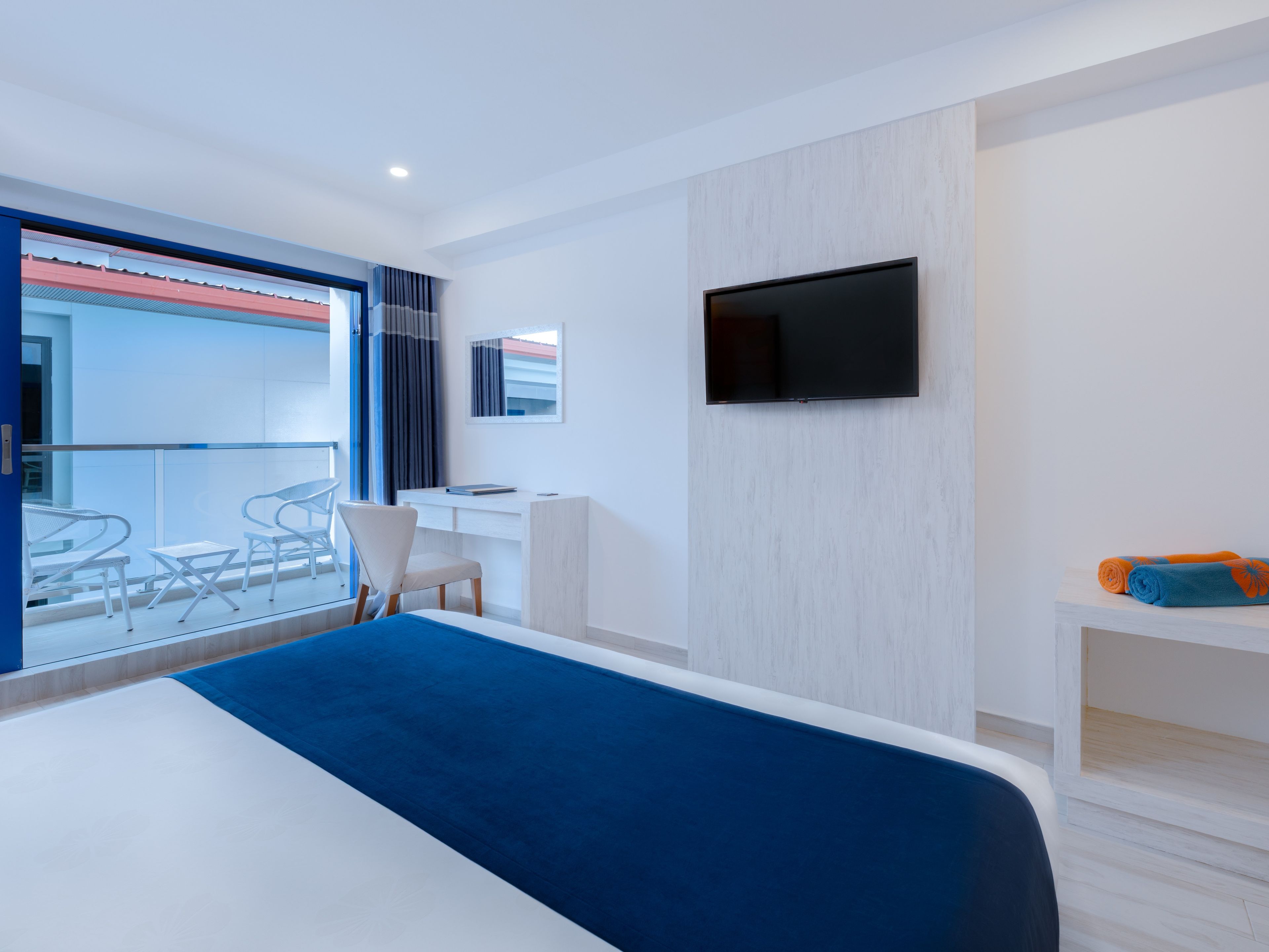 A modern hotel room with a large bed, a desk, a chair, a television, and a balcony with a view.