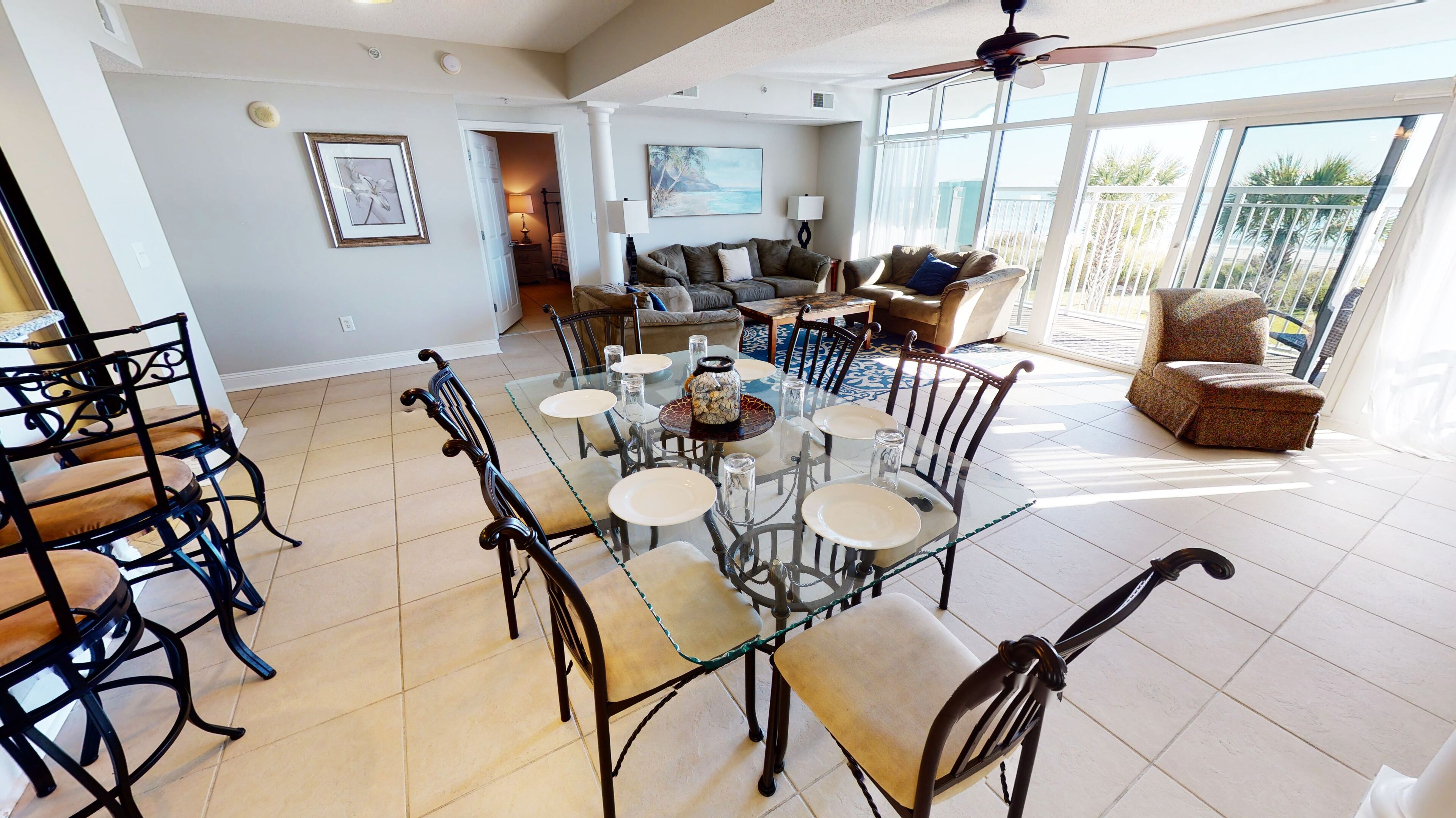 A spacious dining area with a glass table and chairs, a ceiling fan, and a view of the outdoors through large windows.