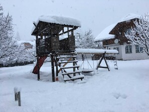 A wooden playhouse with a slide, surrounded by snow-covered trees and a house in the background.