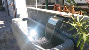 A stone water feature with a metal spout, a stone bench, and a table with chairs in the background.