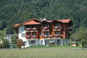 A multi-story hotel with balconies adorned with flowers, set against a forested hillside. - Apartment Stoder - guest house Hartweger (Weißenbach)