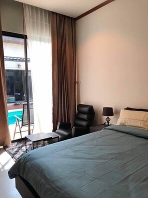 A hotel room with a large bed, a chair, a small table, a lamp, and a view of a swimming pool. - Barbe La Maison (Hua Hin)