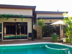 A pool area with a tiled pool, a covered patio with outdoor furniture, and a building with sliding glass doors. - Barbe La Maison (Hua Hin)