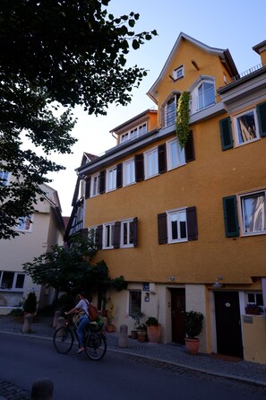 Un immeuble de deux étages avec une façade jaune, de nombreuses fenêtres dotées de volets et un balcon orné d’une plante.