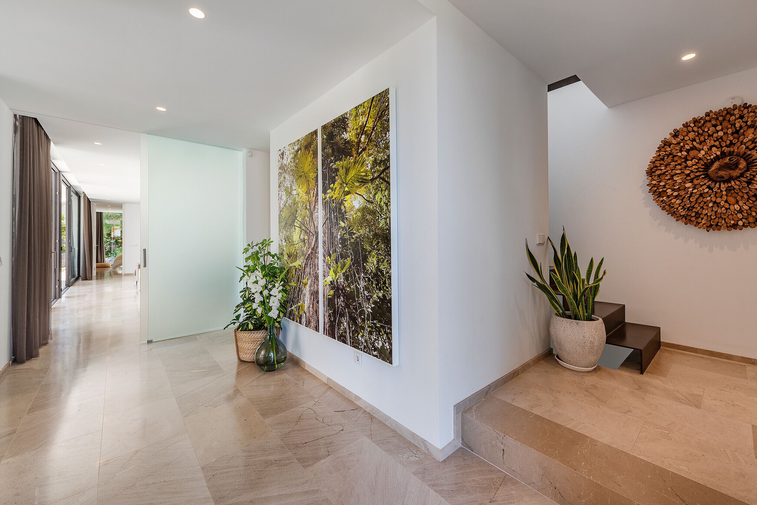 A modern hallway with a wooden floor, a large painting of trees, a potted plant, and a unique wooden wall decoration.