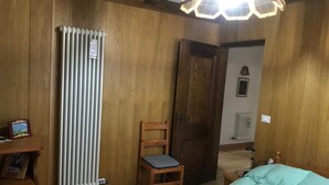 Ein Zimmer mit Holzwänden, einem Heizkörper, einem Bett mit grüner Decke, einem Holzstuhl und einer Tür.