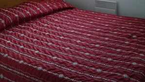 Un dormitorio con cabecera de madera, colcha estampada roja, ventana con cortinas, lámpara de pared y radiador.