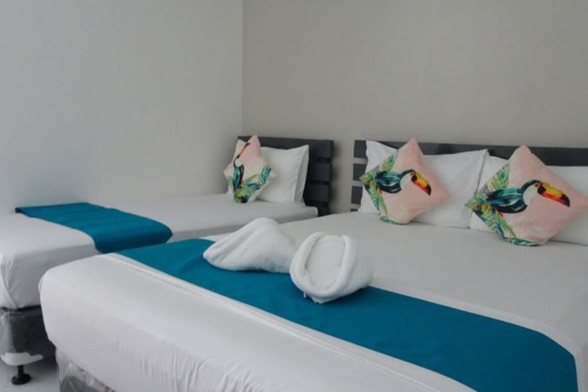 A hotel room with two beds, each with white bedding and teal accents. There are decorative pillows with tropical leaf patterns on the headboards.