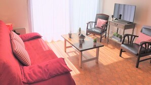 Una sala de estar con un sofá rojo, dos sillones, una mesa de centro, un televisor y una ventana con cortinas traslúcidas.