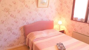Un dormitorio con papel tapiz floral, una cama rosa, una mesita de noche con una lámpara y una ventana con vistas.