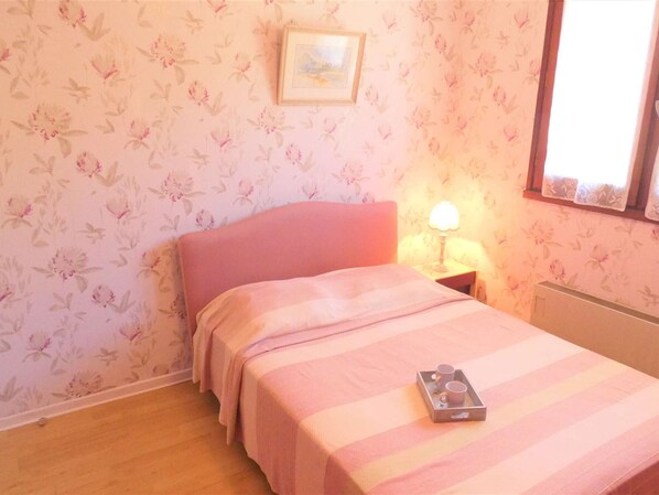 A bedroom with floral wallpaper, a pink bed, a bedside table with a lamp, and a window with a view.