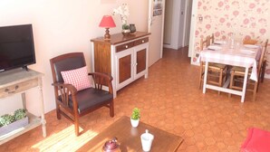Una sala de estar con televisión, un mueble de madera, una silla con cojín a cuadros, una mesa de comedor preparada para dos personas y una planta pequeña sobre una mesa.