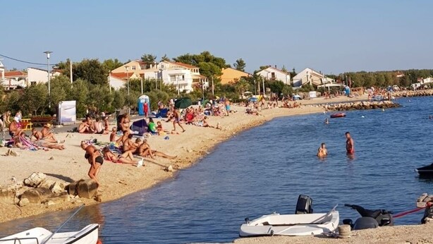 Une plage où l’on voit des gens se prélasser au soleil et nager, des bateaux sur le rivage et des maisons en arrière-plan.