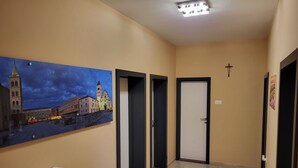 Un couloir aux murs de couleur beige, orné d’un tableau représentant un paysage urbain et d’une croix accrochée au mur.