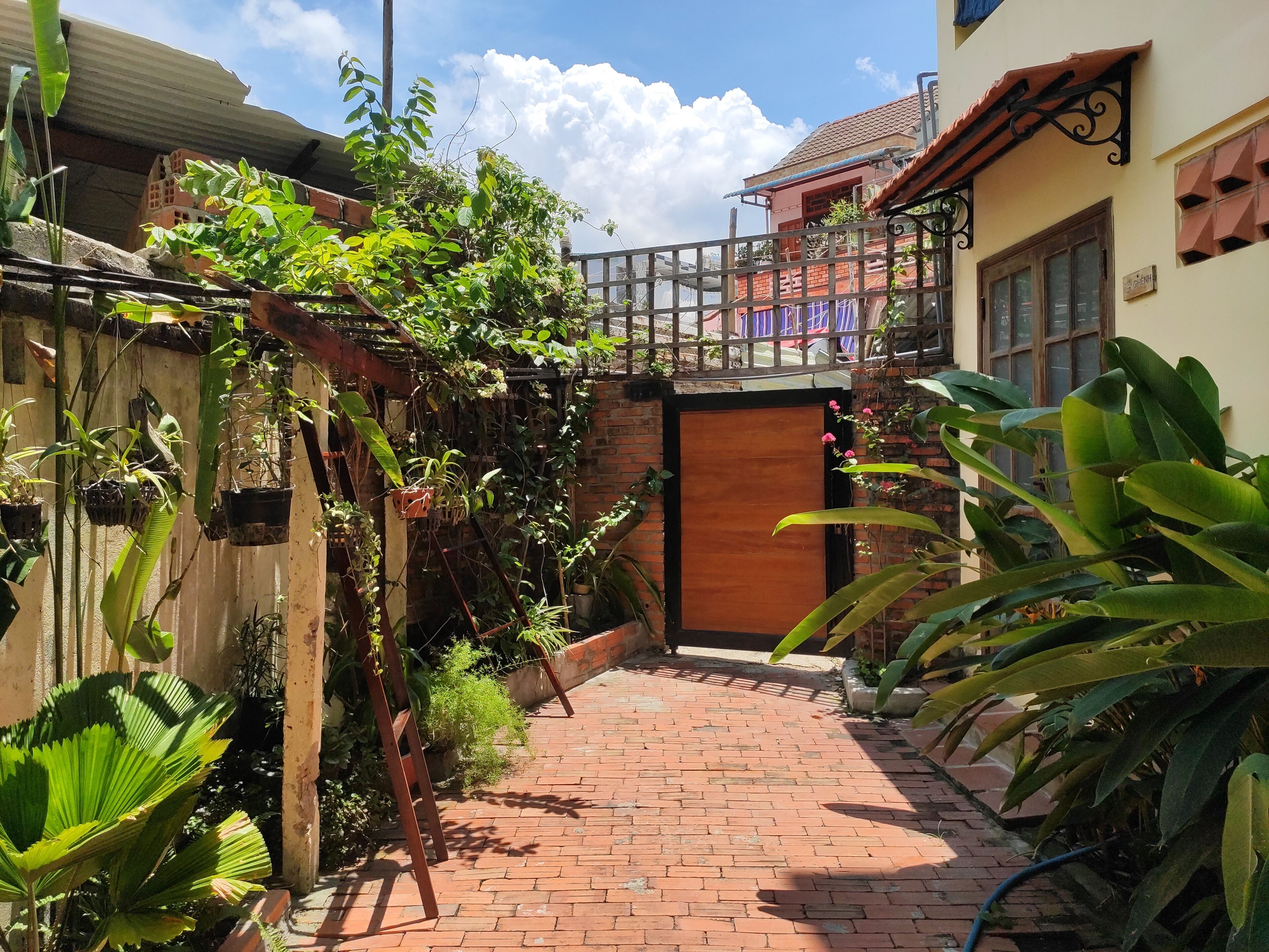 A brick pathway leading to a wooden door, surrounded by lush greenery and a metal fence.