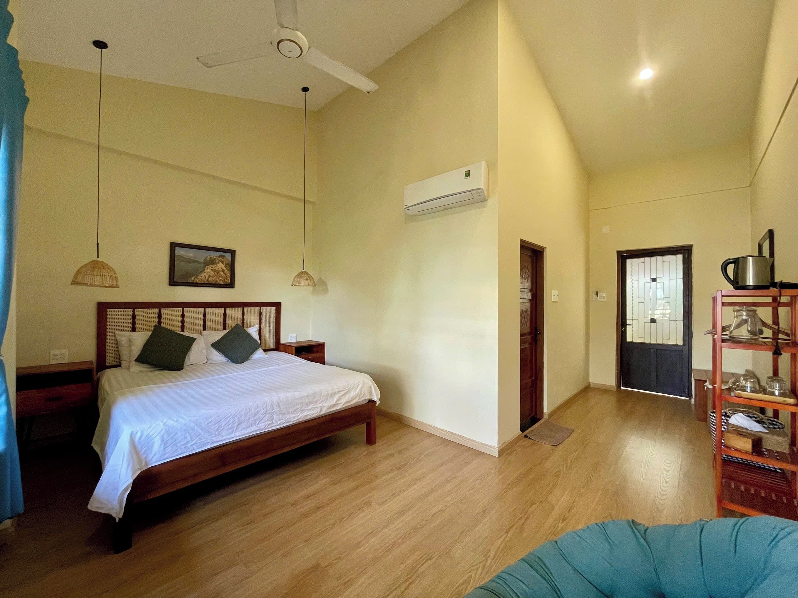 Un dormitorio con cama de madera, dos lámparas de noche, ventilador de techo, equipo de aire acondicionado y una puerta que da a otra habitación.