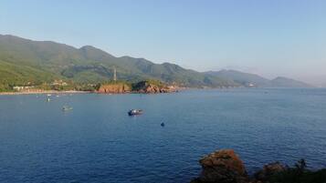 Vista costera con barcos en el agua, un afloramiento rocoso en primer plano y montañas al fondo.