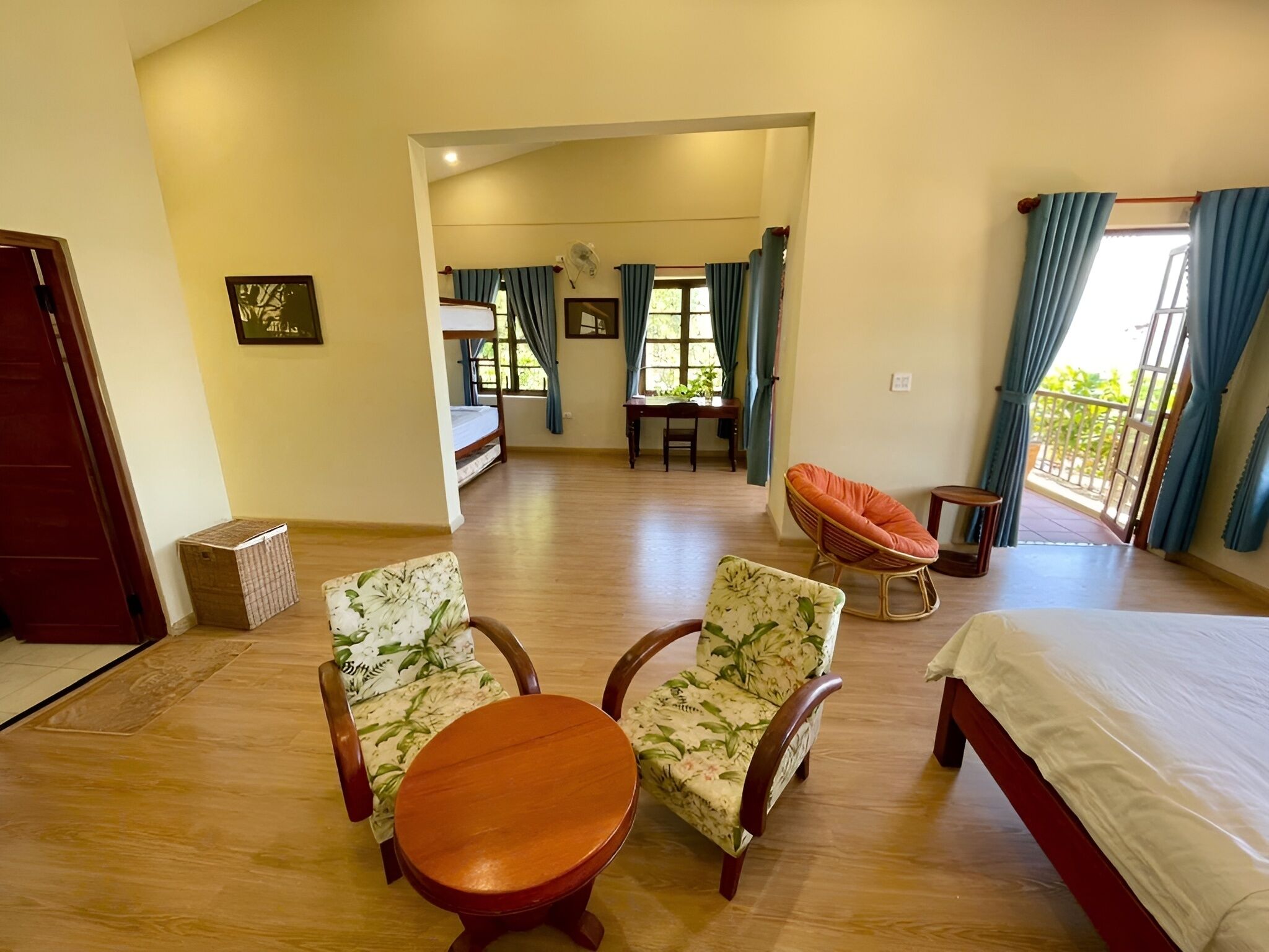 A spacious room with a wooden floor, a bed, two floral-patterned chairs, a small round table, a wicker chair, and a balcony with curtains.