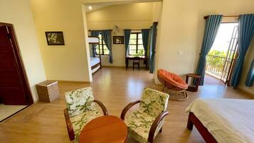 A spacious room with a wooden floor, a bed, two floral-patterned chairs, a small round table, a wicker chair, and a balcony with curtains.